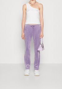 Bílé jednoramenné tričko s kamínkovými detaily doplněné světle fialovými, zamatovými tepláky s elastickým pasem a bočními švy. Bílá sneakers a malá levandulová kabelka dotvářejí celkový vzhled.