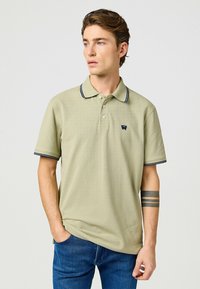 Kaki pikétröja med korta ärmar, marinblå randig krage och ärmslut, med en liten logga på vänster bröst. Bärs med blå jeans.