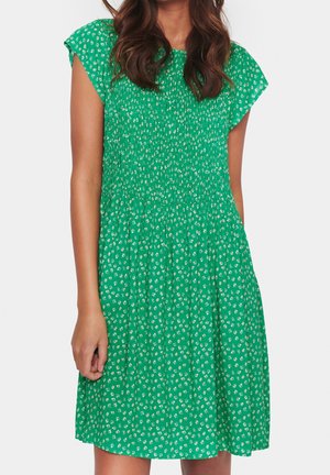 Femme portant une robe verte mi-longue à imprimé floral blanc, manches courtes et tissu froncé sur la poitrine.