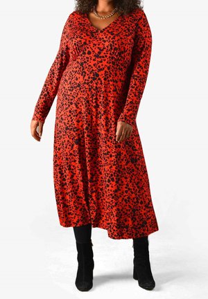 Vrouw draagt een rode midi-jurk met lange mouwen en zwarte abstracte vlekken, zwarte legging, zwarte enkellaarzen en een gouden ketting.