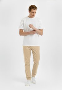 Hvid bomulds T-shirt, rund hals, korte ærmer; beige chinos, slim fit; hvide sneakers; model der står og justerer ur med en neutral baggrund.
