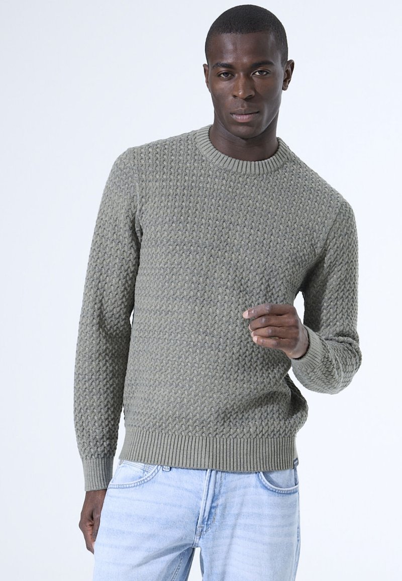 Olivgrüner gestrickter Pullover mit einer strukturierten Musterung, rundem Halsausschnitt und gerippten Bündchen und Saum. Kombiniert mit hellblauen Jeans.