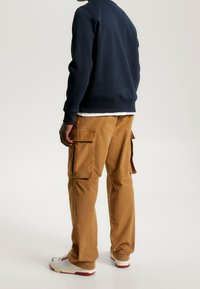 Persona de pie con pantalones cargo marrones, sudadera azul marino y zapatillas blancas con suelas rojas, contra un fondo claro y liso.