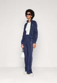 Navy tracksuit with white side stripes, featuring a zip-up jacket and matching pants. Paired with a white top and small handbag.