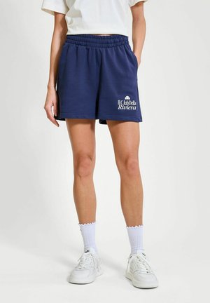 Persona que lleva pantalones cortos con cintura elástica azul marino con el logo "Il Club Della Riviera", zapatillas blancas, calcetines blancos y una camiseta blanca corta.