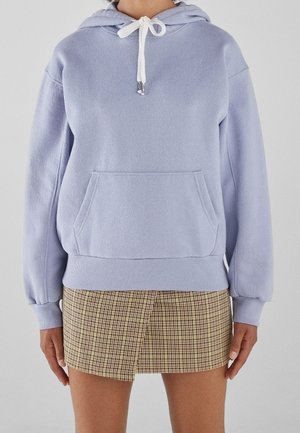 Femme portant un sweat à capuche violet clair avec des cordons blancs et une mini-jupe à carreaux beige sur un fond uni.