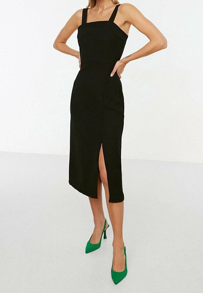 Femme portant une robe midi noire ajustée avec de larges bretelles épaisses et une fente avant, associée à des chaussures à talons verts.