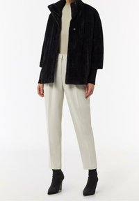 Persoon in een zwarte pluizige jas, beige top, crèmekleurige op maat gemaakte broek en zwarte hoge enkellaarsjes, staand tegen een witte achtergrond.