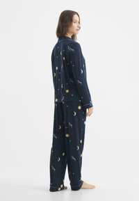 Laivastonsininen pyjamapuku, joka on valmistettu pehmeästä kankaasta ja jossa on leikkisiä kuvioita kuukausista, auringoista ja eläimistä. Sisältää pitkähihaisen paidan ja housut.