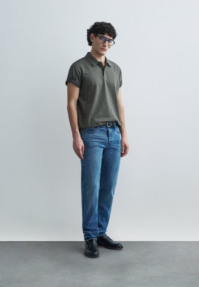 Polo à manches courtes vert olive avec un design texturé, associé à un jean bleu et des chaussures noires. Le polo possède un col classique et des manches retroussées.