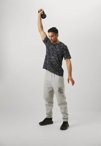 Zwart T-shirt met camouflagemotief en grijze sweatpants met UA-logo. Persoon tilt een kettlebell op en draagt zwarte schoenen. Eenvoudig, sportief ontwerp.