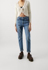 Žena nosící krémový krátký žebrovaný kardigan, modré džíny s vysokým pasem a ohrnutými lemy, tmavé ponožky a masivní hnědé boty na platformě.