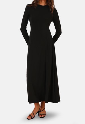 Femme portant une longue robe noire à manches longues et des sandales noires à talons avec des lanières, debout devant un fond clair uni.
