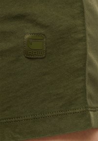 Tissu vert foncé avec ourlet cousu présentant un petit logo carré avec un "G" stylisé et les lettres "RAW."