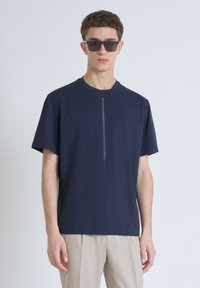 T-shirt navy a maniche corte realizzato in materiale morbido, con un dettaglio a striscia verticale centrale. Indossato con pantaloni chiari e occhiali da sole.