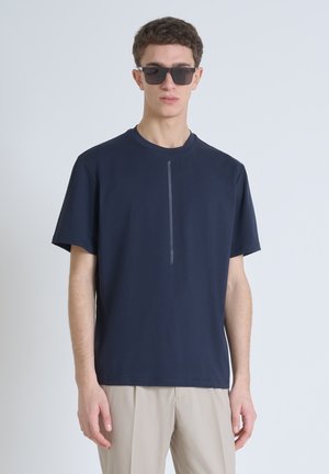 Camiseta navy de manga corta confeccionada en material suave, con un detalle de franja vertical central. Llevada con pantalones de color claro y gafas de sol.
