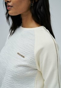 Jersey texturizado en crema con un frente tejido, mangas lisas y acentos metálicos. Presenta una placa con el nombre de la marca en el frente.