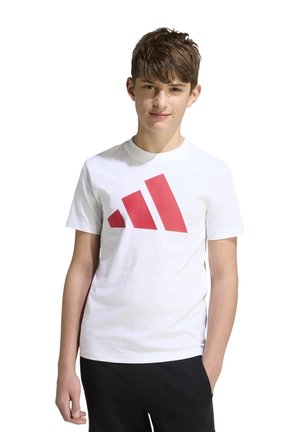Un jeune homme pose sur un fond blanc, vêtu d’un tee-shirt blanc avec un logo Adidas rouge et d’un pantalon noir.