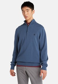 Pullover blu in maglia con zip a un quarto, con polsini e orlo a costine. Presenta un piccolo logo scuro sul petto. Indossato sopra una camicia a collo bordeaux.
