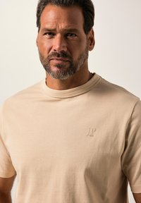 Beige tričko s krátkým rukávem vyrobené z jemné tkaniny, s kulatým výstřihem a reliéfním detailem loga na hrudi. Hladká textura.