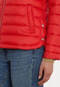 Rode pufferjack die beschikt over een gestikte textuur, zijzak, zilverkleurige hardware en een ronde zoom, gecombineerd met blauwe denim jeans.