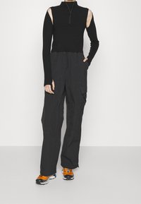 Schwarzes Langarmoberteil mit ausgeschnittenen Schultern, kombiniert mit dunklen Cargo-Hosen; das Outfit verfügt über einen Reißverschluss am Kragen und texturierten Stoff.