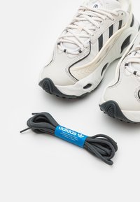 Beige Sportschuhe mit schwarzen Streifen, strukturiertem Obermaterial und Gummisohlen. Ein Paar dunkelgraue Schnürsenkel mit einem blauen Adidas-Tag ist enthalten.