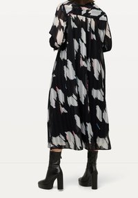 Schwarz-weiß plissiertes Kleid mit abstraktem Muster, transparentem Stoff, weiten Ärmeln und lockerem Schnitt, kombiniert mit schwarzen Plateaustiefeln.