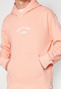 Pfirsichfarbener Kapuzenpullover mit weißem "PORTLAND"-Druck, Kängurutasche, gerippten Bündchen und weicher Textur. Klassisches, lässiges Design.