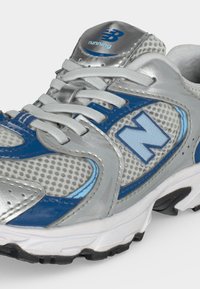 Γκρι και μπλε αθλητικό παπούτσι New Balance με λευκά κορδόνια και αεριζόμενο υφασμάτινο διχτυωτό υλικό, εμφανιζόμενο σε γωνία σε λευκό φόντο.