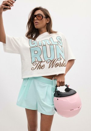Femme portant de grandes lunettes de soleil, un t-shirt blanc court avec le texte « Girls Run The World », un short bleu clair, tenant un casque rose et un téléphone.
