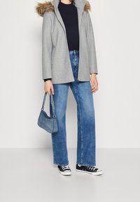 Abrigo gris de lana con capucha de piel sintética, suéter azul marino, jeans azules de pierna ancha y zapatillas negras. Pequeño bolso azul con correa.
