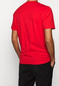 Mand iført en ensfarvet rød t-shirt med korte ærmer og sorte bukser, der står med hænderne i lommerne og vender bort fra kameraet.