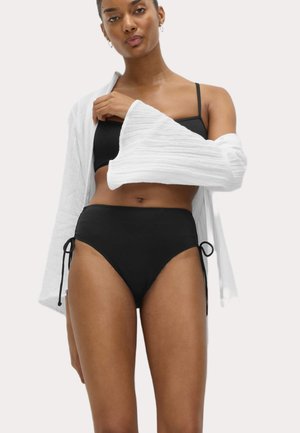 Kvinde iført sort bikini med højtaljede underdele og en let hvid langærmet skjorte hængende ned fra den ene skulder.