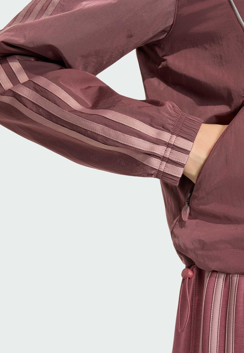 Main dans la poche d'une veste coupe-vent mauve avec poignets élastiques et pantalon mauve assorti avec des détails rayés.