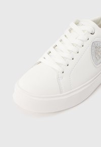 Bijela tenisica s glatkim sintetičkim gornjim dijelom, dizajnom na vezanje i teksturiranim gumeniim potplatom. Ima detalj sa sjajnim elementom sa strane.