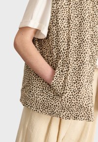 Beige leopardmönstrad väst med två sidofickor, tillverkad av mjukt tyg. Designen har en subtil textur och en lös passform, ihop med en vit topp.