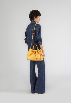 Gele leren handtas met gerimpelde opening, korte handvatten en verstelbare schouderriem, gedragen door een persoon in een denim jumpsuit, met de rug naar de camera gekeerd.