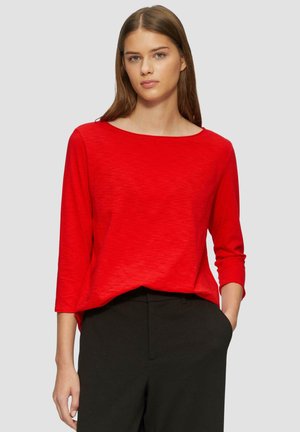Haut rouge à manches longues avec un col bateau, fabriqué en matériau léger. Présente une coupe décontractée et un design simple, associé à un pantalon sombre.