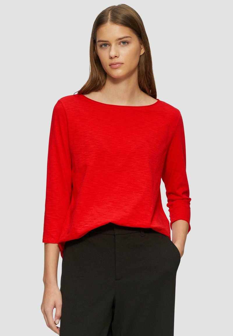 Haut rouge à manches longues avec un col bateau, fabriqué en matériau léger. Présente une coupe décontractée et un design simple, associé à un pantalon sombre.