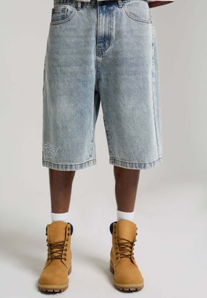 Mężczyzna ubrany w jasnoniebieskie dżinsowe spodenki do kolan i jasnobrązowe, sznurowane buty robocze z białymi skarpetkami na neutralnym tle.