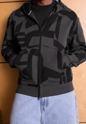 Person trägt einen schwarz-grau mit abstraktem Muster bedruckten Hoodie mit Reißverschluss, die Hände in den Taschen, hellblaue Jeans vor einem Holzpaneel-Hintergrund.