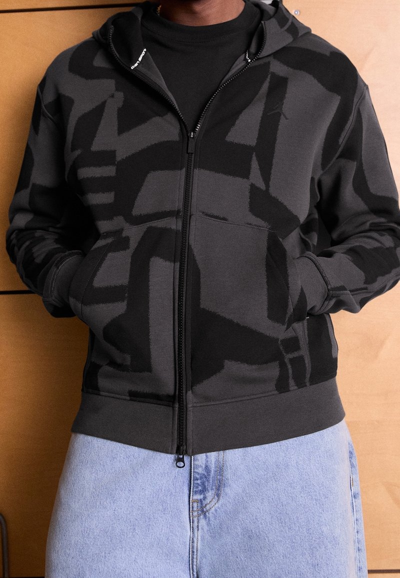 Personne portant un sweat à capuche noir et gris à motifs abstraits avec fermeture éclair, mains dans les poches et jean en denim bleu clair, devant un fond en panneaux de bois.