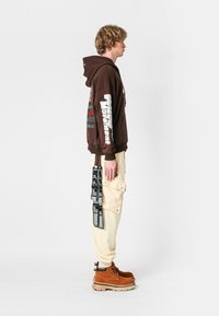 Joven de pie de lado, vistiendo una sudadera marrón con texto blanco en las mangas, pantalones cargo crema con detalles de correa negra y botas color arena.