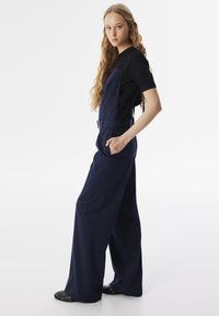 Salopette blu navy con una vestibilità comoda, caratterizzata da gambe larghe, spalline regolabili e una tasca frontale. Abbinata a una maglietta nera a maniche corte.