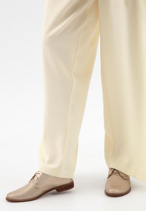 Beige leren veterschoenen met perforaties, gedragen met loszittende crèmekleurige broeken tegen een lichte achtergrond.