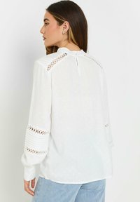 Blusa bianca con un motivo testurizzato, caratterizzata da maniche lunghe con dettagli in pizzo e chiusura a occhio di bue sul retro. Tessuto morbido e leggero.