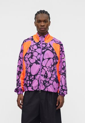 Jeune homme aux cheveux tressés portant une veste à motifs violets et noirs avec des accents en mesh orange vif et un pantalon noir, debout sur un fond blanc.