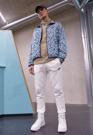 Blaue und weiße floral gemusterte Bomberjacke über einem beigen Hemd, kombiniert mit weißen Jogginghosen und weißen High-Top-Sneakers.