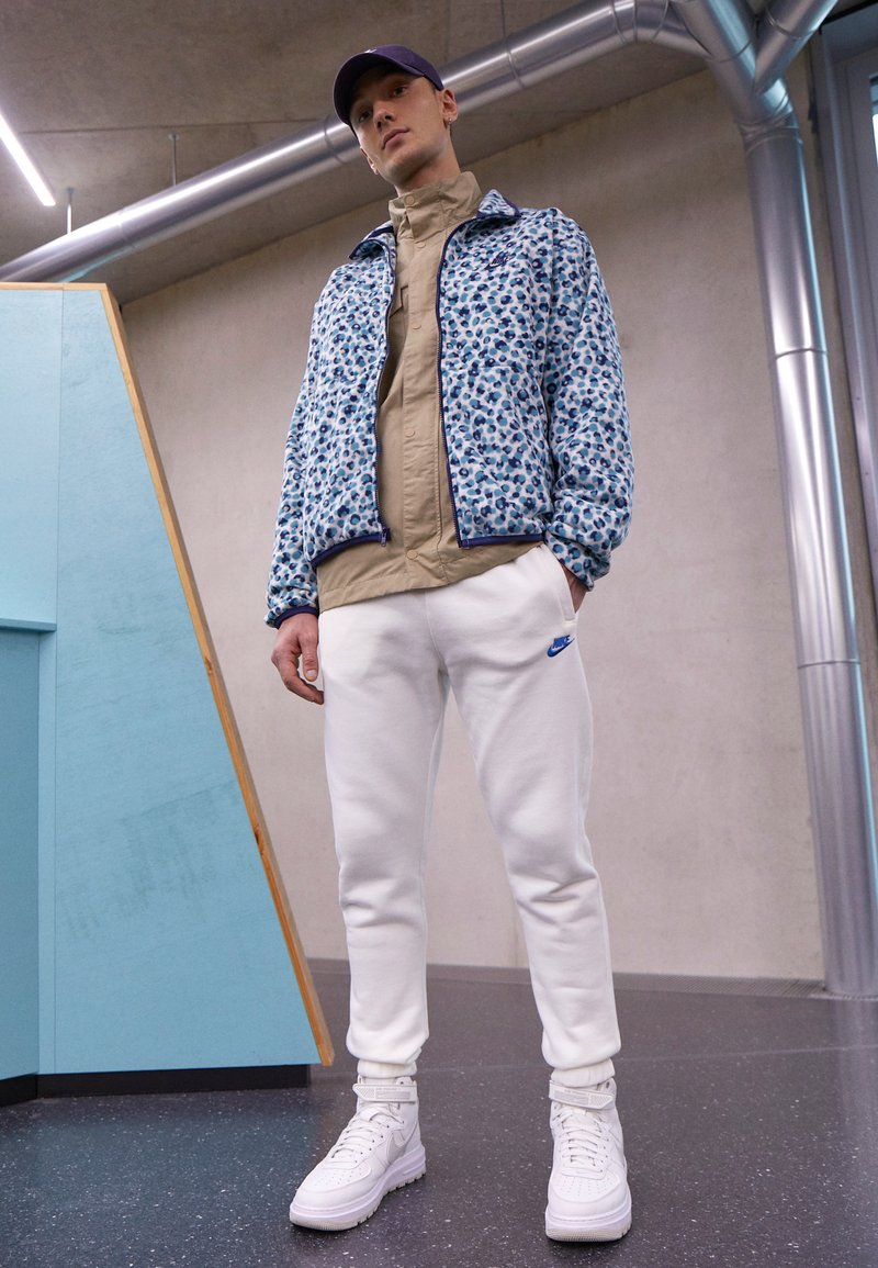 Jachetă bomber cu un model floral albastru și alb, purtată peste o cămașă bej, asortată cu pantaloni de trening albi și adidași înalți albi.